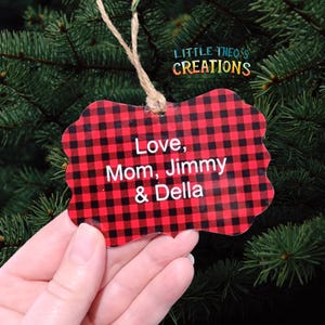 Puede incluir: Adorno navide&ntilde;o a cuadros rojos y negros con el texto "Love, Mom, Jimmy & Della". El adorno est&aacute; colgado de una cuerda contra un fondo de agujas de pino verdes. El adorno tiene un borde decorativo.