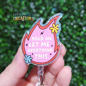 May include: A pink, flame-shaped badge reel with the text "HOLD ON, LET ME OVERTHINK THIS." The badge reel has a glittery blue border and features flower and lightning bolt graphics. The brand name "LITTLE THEO'S CREATIONS" is at the top.