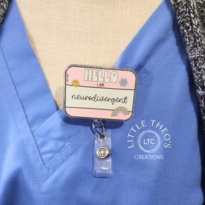 Könnte beinhalten: Rosa glitzernder Badgehalter mit dem Text "Hello I am neurodivergent" mit einem Smiley, einer Blume und einem Regenbogen. Der Badgehalter ist an einer silbernen, einziehbaren Badge-Spule mit einem durchsichtigen Kunststoffgehäuse befestigt. Der Badgehalter trägt den Text "Little Theo's LTC Creations" unten.