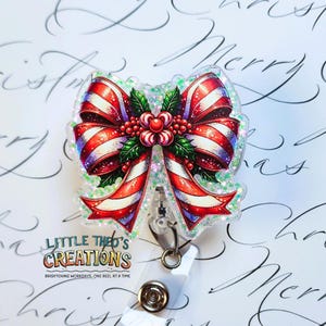 May include: A festive badge reel featuring a Christmas bow design with red and white stripes, holly leaves, and berries. The badge reel has a clear plastic base and a retractable cord. The text "Little Theo's Creations" is visible.