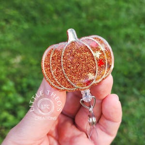 May include: A pumpkin-shaped badge reel featuring a vibrant orange glitter finish with embedded gold and red leaf-shaped accents. The design is outlined in a metallic silver tone and includes a clear plastic retractable cord and a metal clip.
