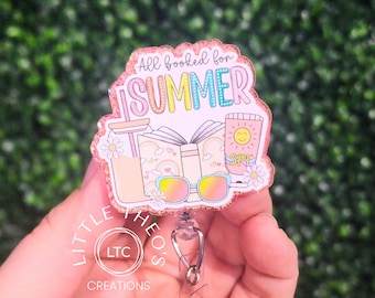 Puede incluir: Un portatarjetas con temática de verano. La insignia presenta el texto "All booked for SUMMER" con ilustraciones de una bebida, un libro, gafas de sol y protector solar. La insignia es rosa y brillante, con un carrete retráctil blanco y un clip de metal.