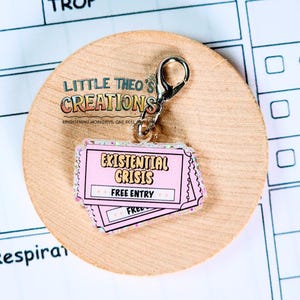 May include: A pink and glittery keychain charm shaped like two tickets, with the words "Existential Crisis" and "Free Entry" printed on them. The charm is attached to a silver clasp and sits on a wooden circle. The text "Little Theo's Creations" is also visible.