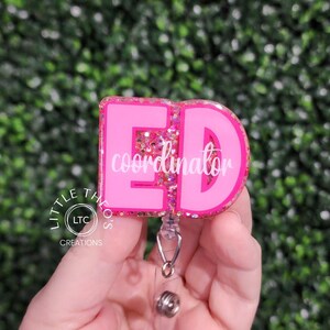 May include: A pink badge reel with the letters "ED" in a bold font, with the word "coordinator" in white script. The badge reel has a glittery border and a retractable cord with a metal clip. The badge is held in a hand.