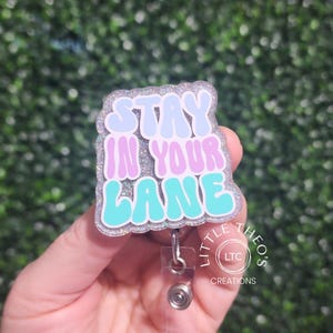 Könnte beinhalten: Ein silberner Glitzer-Badgehalter mit dem Text "Stay in your lane" in Blau, Rosa und Türkis. Der Badgehalter hat eine silberne, einziehbare Badge-Rolle.