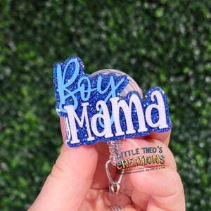 May include: A blue glitter badge reel with the words "Boy Mama" in a white and blue design. The badge reel also has the text "LITTLE THEO'S CREATIONS" and the tagline "BRIGHTENING WORKDAYS, ONE REEL AT A TIME".