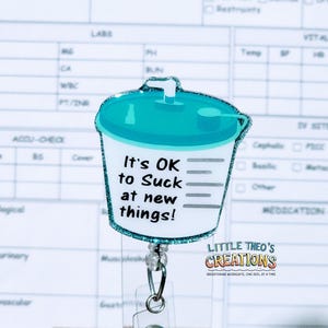May include: A teal and white badge reel with a glittery border, shaped like a medical suction canister. The text reads "It's OK to Suck at new things!" The reel includes a clear plastic ID holder and a metal clip. The logo "LITTLE THEO'S CREATIONS" is also visible.