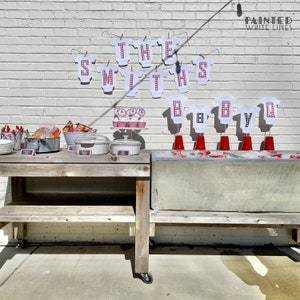 Op de afbeelding: Een babyborrel-opstelling met een houten tafel en een metalen koelbox. De tafel is versierd met rood-wit geruit papier en heeft een bord met de tekst "The Smiths Baby BBQ".