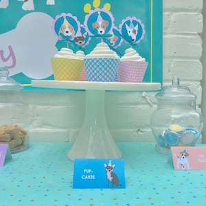 May include: A white cake stand with four cupcakes decorated with dog faces and party hats. The cupcakes are on a blue and white polka dot tablecloth. A sign reads "Pup-Cakes".