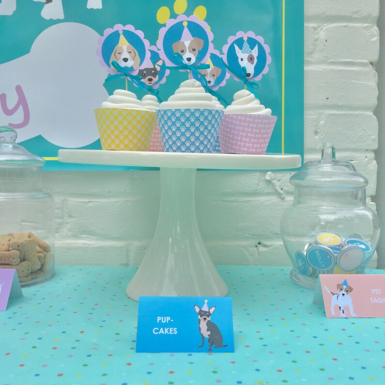 May include: A white cake stand with four cupcakes decorated with dog faces and party hats. The cupcakes are on a blue and white polka dot tablecloth. A sign reads "Pup-Cakes".