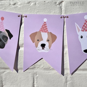 May include: A purple banner with three dog faces wearing pink party hats. The dogs are a pug, a bulldog, and a white dog. The banner is decorated with a red and white string.