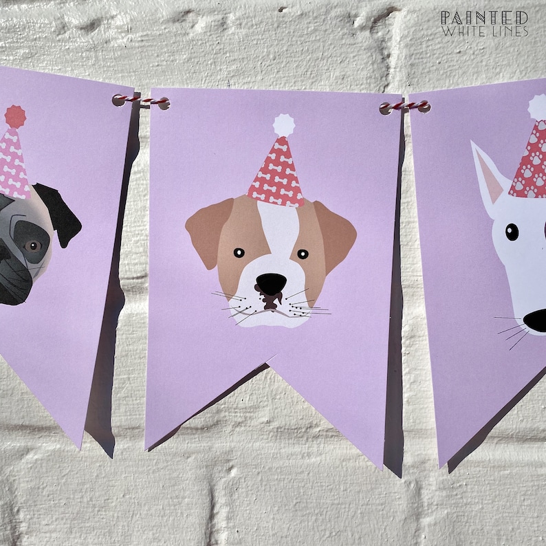 May include: A purple banner with three dog faces wearing pink party hats. The dogs are a pug, a bulldog, and a white dog. The banner is decorated with a red and white string.
