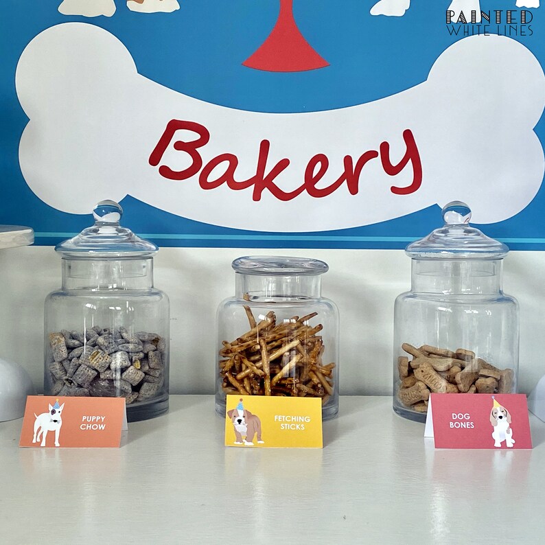May include: Three glass jars filled with dog treats. The jars are labeled "Puppy Chow", "Fetching Sticks", and "Dog Bones". The jars are on a white counter with a blue and white dog bone sign above them that says "Bakery".