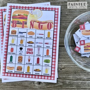 May include: A red and white checkered bingo card with a hamburger and hot dog design. The card has a free space in the middle and is filled with various food and grilling related items. The card is on a wooden table with a glass bowl of bingo chips.