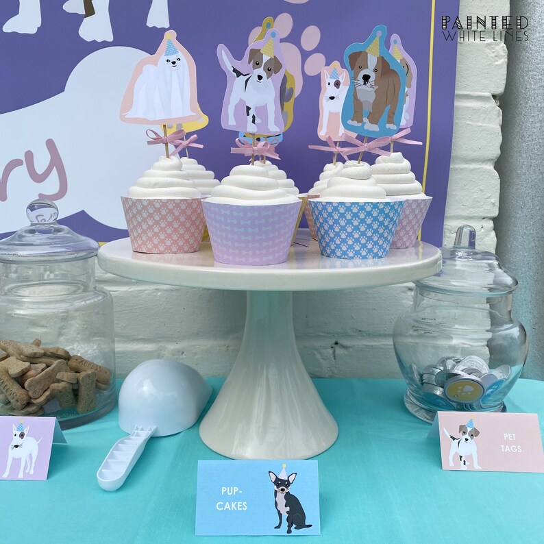 Puede incluir: Un soporte para pasteles blanco con tres cupcakes decorados con adornos de perros. Los cupcakes est&aacute;n sobre una superficie azul claro con un cartel que dice "Pup-Cakes". Tambi&eacute;n hay favores de fiesta con tem&aacute;tica de perros en un frasco de vidrio y un cartel que dice "Pet Tags".