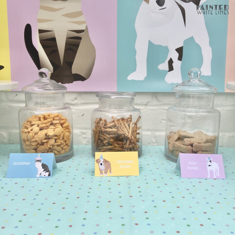 May include: Three glass jars filled with dog treats on a blue and white polka dot tablecloth. The jars are labeled "Goldfish", "Fetching Sticks", and "Dog Bones". Each label has a cartoon dog or cat wearing a party hat.