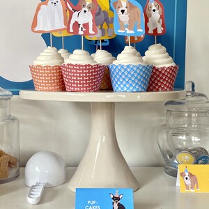 May include: A white cake stand with five cupcakes decorated with dog toppers. The cupcakes are topped with white frosting and have different colored wrappers. The toppers are of different dog breeds wearing party hats. The stand is on a white surface with a sign that says "Pup-Cakes".