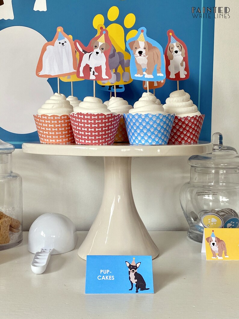 May include: A white cake stand with five cupcakes decorated with dog toppers. The cupcakes are topped with white frosting and have different colored wrappers. The toppers are of different dog breeds wearing party hats. The stand is on a white surface with a sign that says "Pup-Cakes".