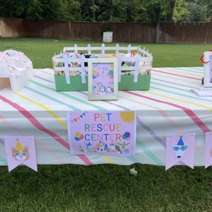 May include: A pink and white striped tablecloth with a sign that says "Pet Rescue Center" and a white picket fence with stuffed animals inside. There are also pink and white banners with cartoon animals on them.