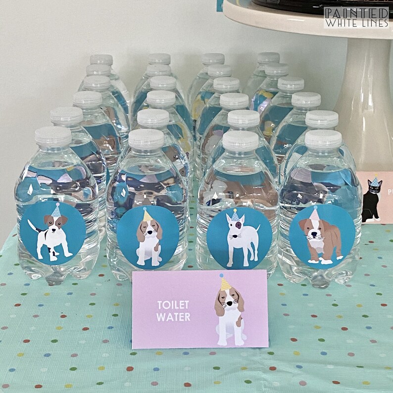 May include: A table with a light blue tablecloth with white polka dots. There are 15 clear plastic water bottles with blue and white dog stickers on them. The stickers feature dogs wearing party hats. A sign on the table reads "Toilet Water".