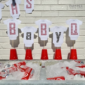 May include: Red and black checkered baby onesies with the letters "BABY Q" on sticks in red solo cups. The letters are on a white brick wall with the words "Painted White Lines" above them. There are cans of soda in a cooler with ice.