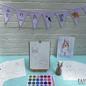 May include: A table set up for a children's art party with a banner featuring animals wearing party hats. There are several coloring pages of animals wearing party hats, a set of watercolors, and paintbrushes. The text "Painted White Lines" is visible in the bottom right corner of the image.