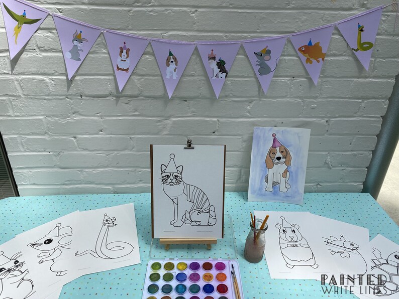 May include: A table set up for a children's art party with a banner featuring animals wearing party hats. There are several coloring pages of animals wearing party hats, a set of watercolors, and paintbrushes. The text "Painted White Lines" is visible in the bottom right corner of the image.