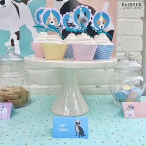 May include: A white cake stand with four cupcakes decorated with dog and cat toppers. The cupcakes are on a blue and white polka dot tablecloth. The text "Pup Cakes" is on a blue sign in front of the cupcakes.