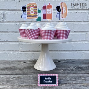 May include: Vanilla cupcakes decorated with red and white checkered cupcake liners and topped with a variety of barbecue themed cupcake toppers, including a hamburger, hot dog, grill, and ketchup and mustard bottles. The cupcakes are displayed on a white cake stand with a sign that reads "Vanilla Cupcakes".