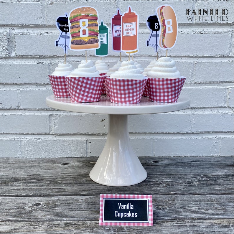 May include: Vanilla cupcakes decorated with red and white checkered cupcake liners and topped with a variety of barbecue themed cupcake toppers, including a hamburger, hot dog, grill, and ketchup and mustard bottles. The cupcakes are displayed on a white cake stand with a sign that reads "Vanilla Cupcakes".