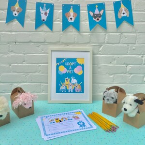 May include: A framed poster with the text "Adopt a Pet" and illustrations of dogs and balloons. There are also stuffed dog toys in brown paper bags, a certificate of adoption, and pencils on a blue and white patterned table.