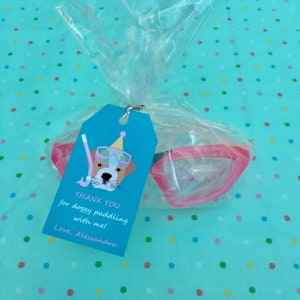 May include: A gift bag containing pink swimming goggles and a blue tag. The tag features a cartoon dog wearing goggles and a party hat, with the text "THANK YOU for doggy paddling with me! Love, Alessandra."