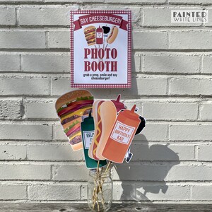 May include: A photo booth sign with a red and white checkered background. The sign says "Say Cheeseburger! Photo Booth grab a prop, smile and say cheeseburger!" There are several props in a glass jar, including a hamburger, hot dog, ketchup bottle, and a sign that says "Happy Birthday Kai!"