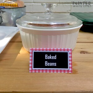 Puede incluir: Una cacerola blanca con tapa de vidrio llena de frijoles horneados. Un letrero a cuadros rojos y blancos dice "Baked Beans".