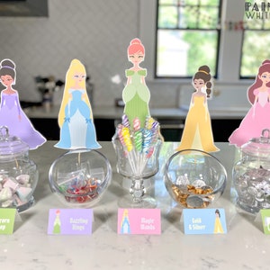 May include: A table setting with five glass jars filled with different candies. Each jar has a princess character cutout and a sign with the name of the candy. The signs read "Unicorn Poop", "Dazzling Rings", "Magic Sands", "Gold & Silver", and "Unicorn Kisses".
