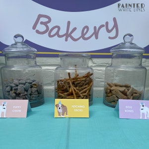 Puede incluir: Tres tarros de vidrio llenos de golosinas para perros etiquetados como "Puppy Chow", "Fetching Sticks" y "Dog Bones". Los tarros est&aacute;n sobre una mesa azul claro con un letrero arriba que dice "Bakery" en letras rosas en cursiva con las palabras "Painted White Lines" en letras blancas sobre la palabra "Bakery".