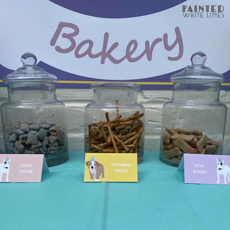 Puede incluir: Tres tarros de vidrio llenos de golosinas para perros etiquetados como "Puppy Chow", "Fetching Sticks" y "Dog Bones". Los tarros est&aacute;n sobre una mesa azul claro con un letrero arriba que dice "Bakery" en letras rosas en cursiva con las palabras "Painted White Lines" en letras blancas sobre la palabra "Bakery".