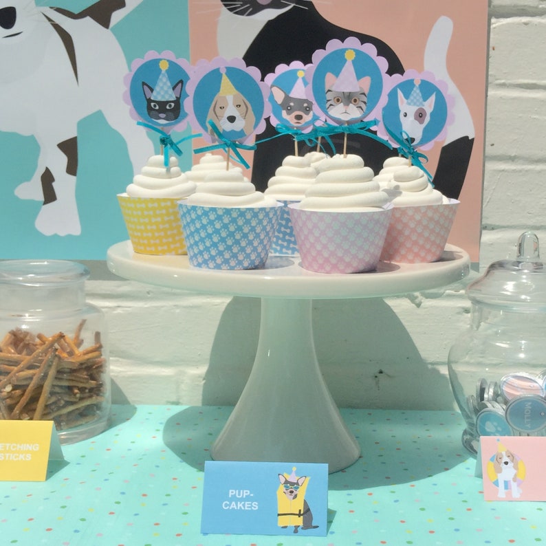 May include: A display of cupcakes with white frosting, each in a patterned liner, topped with dog and cat-themed toppers. The cupcakes sit on a white cake stand, with a sign that reads "Pup-Cakes". The background includes dog and cat illustrations.