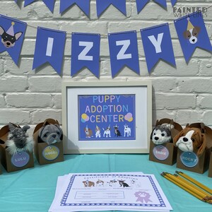 May include: A blue and white banner with the name "IZZY" on it. A framed sign that says "Puppy Adoption Center" with a row of cartoon puppies. Five brown boxes with stuffed puppies inside. Each box has a name tag: Lulu, Angel, Sadie, Rocky. A certificate of adoption with a pink ribbon.