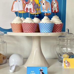 May include: A white cake stand with five cupcakes decorated with dog toppers. The cupcakes are topped with white frosting and have different colored wrappers. The toppers are of different dog breeds wearing party hats. The stand is on a white surface with a sign that says "Pup-Cakes".