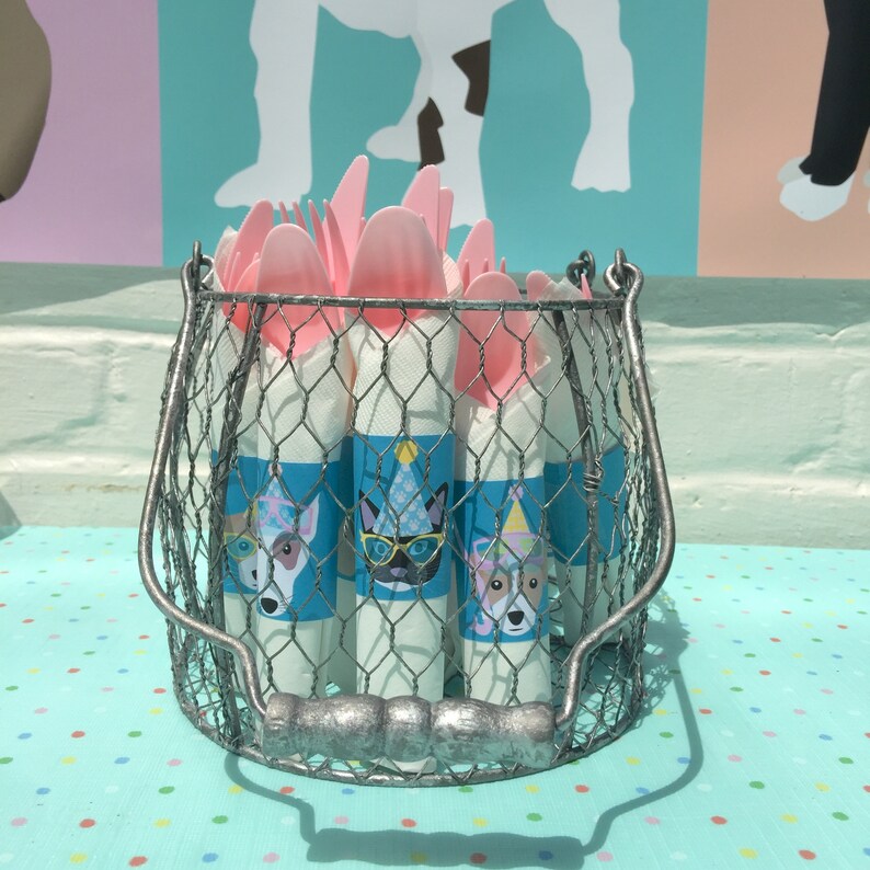 May include: A wire basket filled with pink plastic forks and napkins wrapped in blue and white paper with dog illustrations. The napkins have a blue background with a white border and a white dog wearing a party hat.