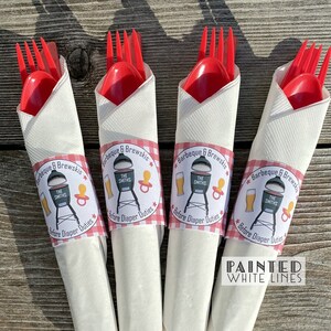 May include: Four white napkins with red and white checkered bands wrapped around them. Each napkin has a round sticker with a grill, beer, and pacifier graphic. The stickers say "Barbeque & Brewskis" and "The Smiths" with the text "Before Diaper Duties" below the graphic. The text "Painted White Lines" is below the napkins.