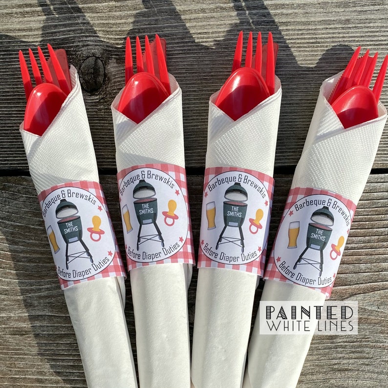 May include: Four white napkins with red and white checkered bands wrapped around them. Each napkin has a round sticker with a grill, beer, and pacifier graphic. The stickers say "Barbeque & Brewskis" and "The Smiths" with the text "Before Diaper Duties" below the graphic. The text "Painted White Lines" is below the napkins.