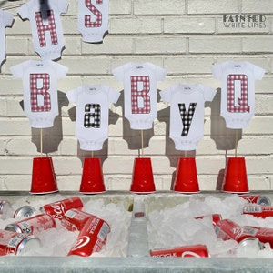 Op de afbeelding: Rode en zwarte geruite baby-rompers met de letters "BABY Q" op stokjes in rode plastic bekers. De letters bevinden zich op een witte bakstenen muur met de tekst "Painted White Lines" erboven. Er zijn blikjes frisdrank in een koeler met ijs.