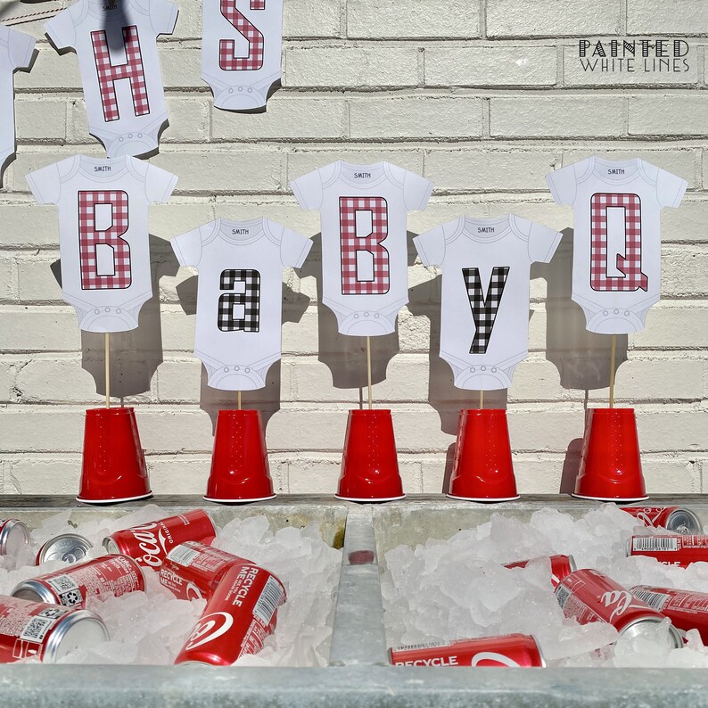 Op de afbeelding: Rode en zwarte geruite baby-rompers met de letters "BABY Q" op stokjes in rode plastic bekers. De letters bevinden zich op een witte bakstenen muur met de tekst "Painted White Lines" erboven. Er zijn blikjes frisdrank in een koeler met ijs.