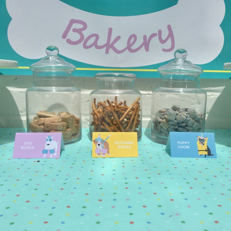 May include: Three glass jars filled with dog treats labeled "Dog Bones", "Fetching Sticks", and "Puppy Chow". The jars are on a blue and white polka dot table cloth with the word "Bakery" in pink letters above them.