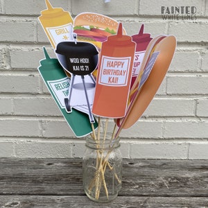 K&ouml;nnte beinhalten: Ein Set aus 7 bunten Papp-Fotobooth-Requisiten mit Grill-Thema. Die Requisiten beinhalten einen Grill, ein Hotdog, einen Hamburger, Ketchup, Senf, Relish und ein Schild mit der Aufschrift "Happy Birthday Kai!"