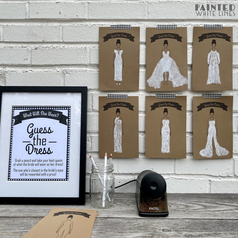 May include: A bridal shower game with a framed sign that says "What Will She Wear? Guess the Dress" and eight cards with different wedding dress styles. The cards are brown with white text and white drawings of the dresses. The game includes a pencil, a pencil sharpener, and a glass jar.