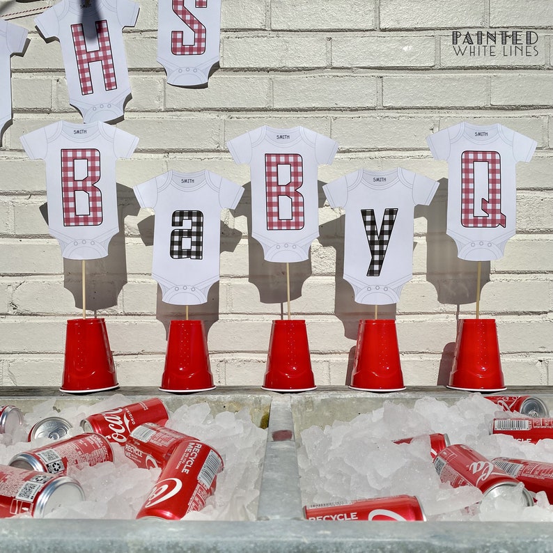 May include: A baby shower party decoration with red and black gingham patterned onesies with the letters "BABY Q" on them. The letters are on sticks and placed in red plastic cups. The cups are on a table with a cooler full of ice and soda cans.