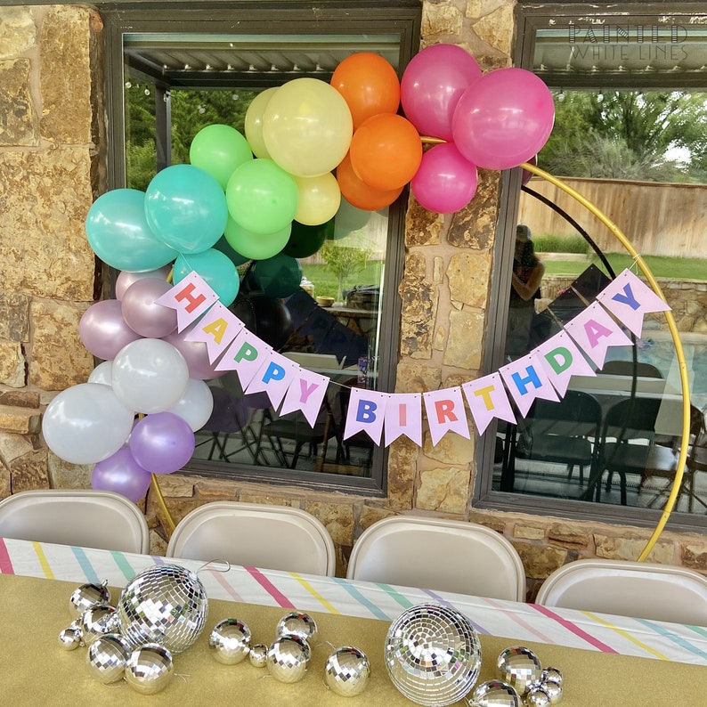 May include: A rainbow balloon arch with a pink banner that reads "Happy Birthday" in colourful letters. The arch is made of balloons in various colours, including pink, purple, white, green, yellow, and orange. The banner is hanging from the arch and is made of pink paper with colourful letters. The arch is set up in front of a window with a brick wall behind it. There are silver disco balls on a table in the foreground.